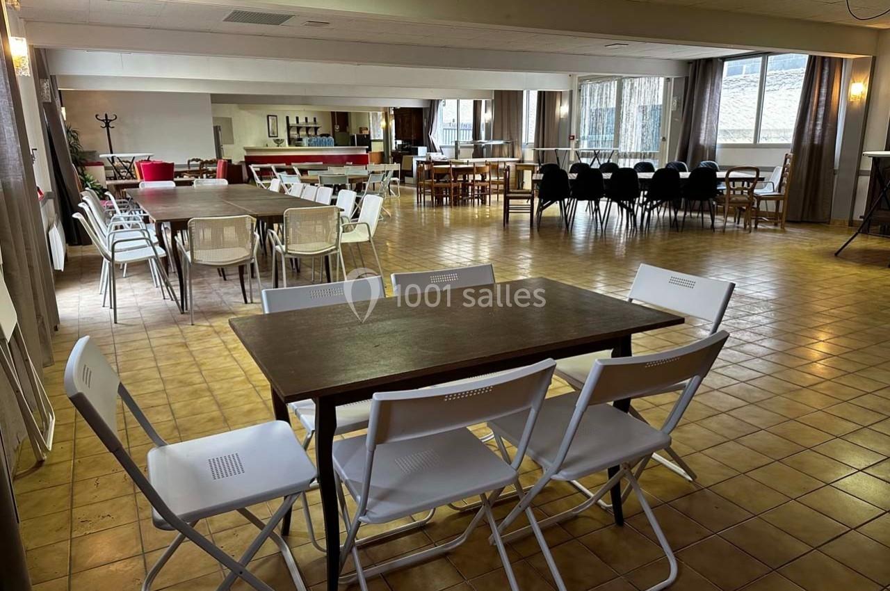 Salle spacieuse avec tables et chaises disposées pour une réunion ou un événement, éclairée par des fenêtres.