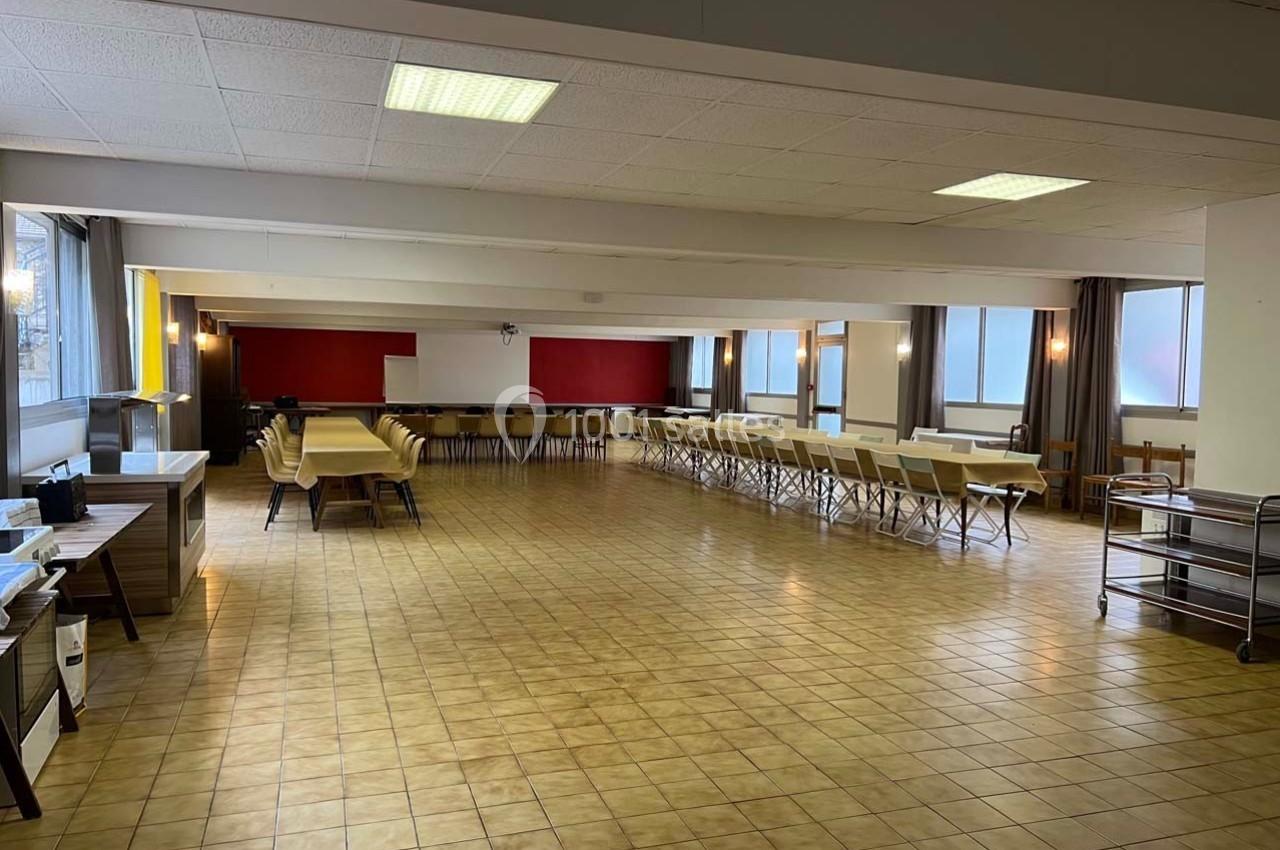Salle spacieuse avec tables et chaises alignées, sol en carrelage beige, murs clairs et fenêtres laissant entrer la lumière.