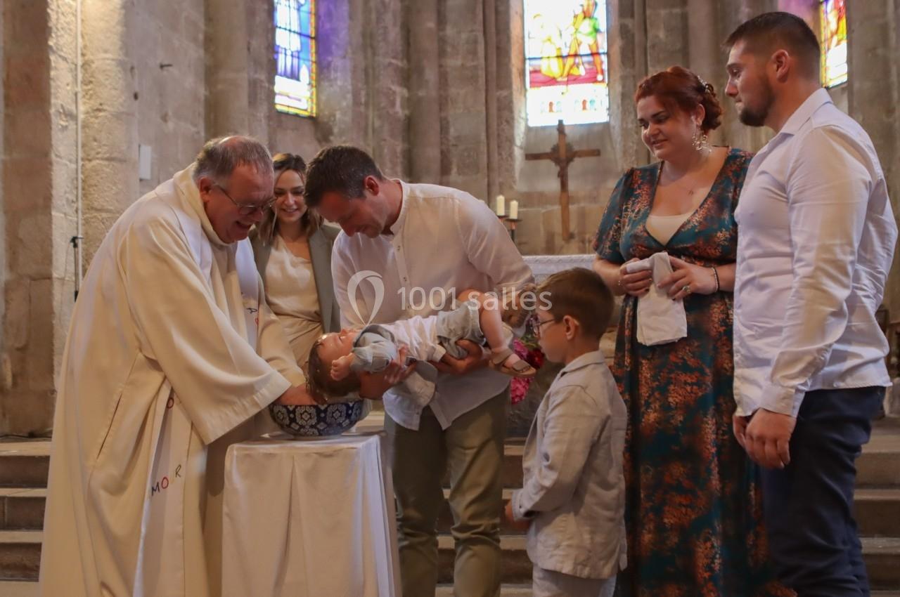 Un prêtre baptise un bébé dans une église, entouré de membres de la famille et de vitraux en arrière-plan.