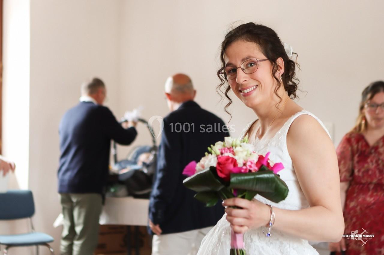 Une femme en robe blanche sourit en tenant un bouquet de fleurs, avec des personnes en arrière-plan flou.