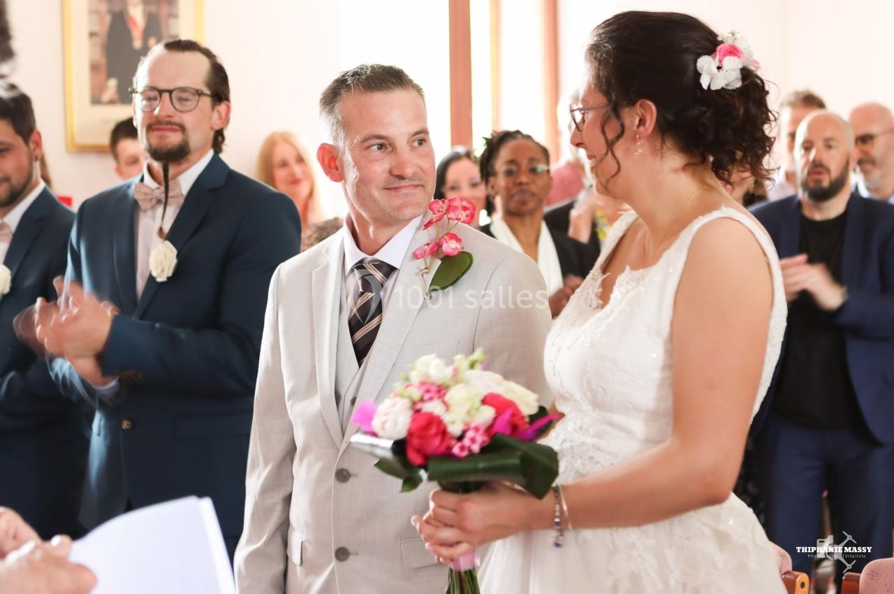 Un couple en tenue de mariage échange un regard complice dans une salle entourée d'invités applaudissant.
