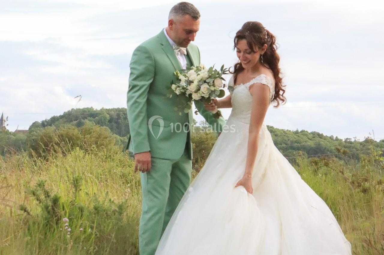 Un couple en tenue de mariage se tient dans un champ verdoyant, la mariée tenant un bouquet de fleurs blanches.