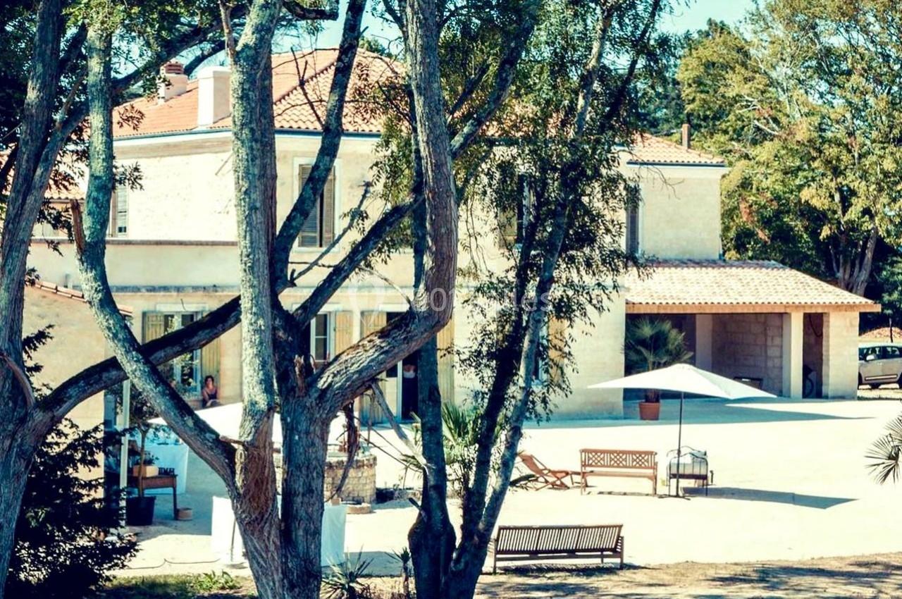 Grande maison en pierre entourée d'arbres, avec une cour ensoleillée, des bancs et un parasol.