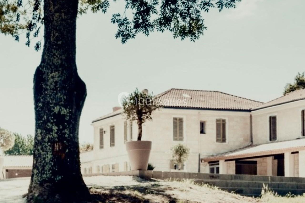 Bâtiment en pierre avec toit en tuiles, entouré d'arbres et d'un mur bas, sous un ciel clair.