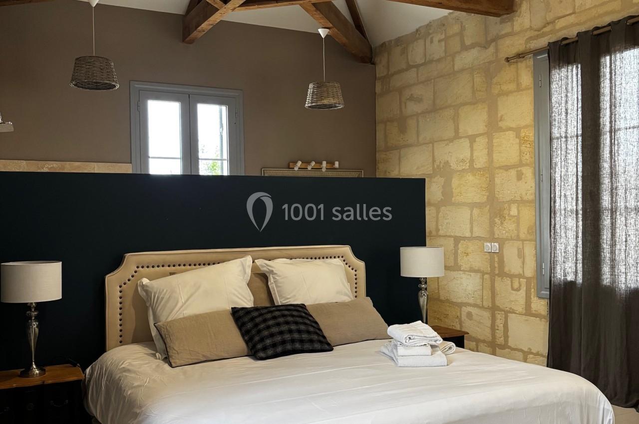 Chambre lumineuse avec lit double, poutres apparentes, murs en pierre et rideaux filtrant la lumière.