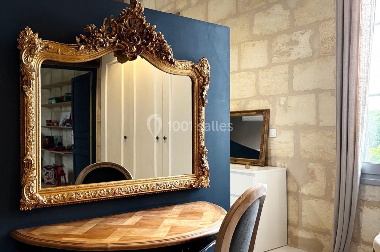 Miroir doré orné au-dessus d'une table en bois, dans une pièce lumineuse aux murs en pierre et poutres apparentes.
