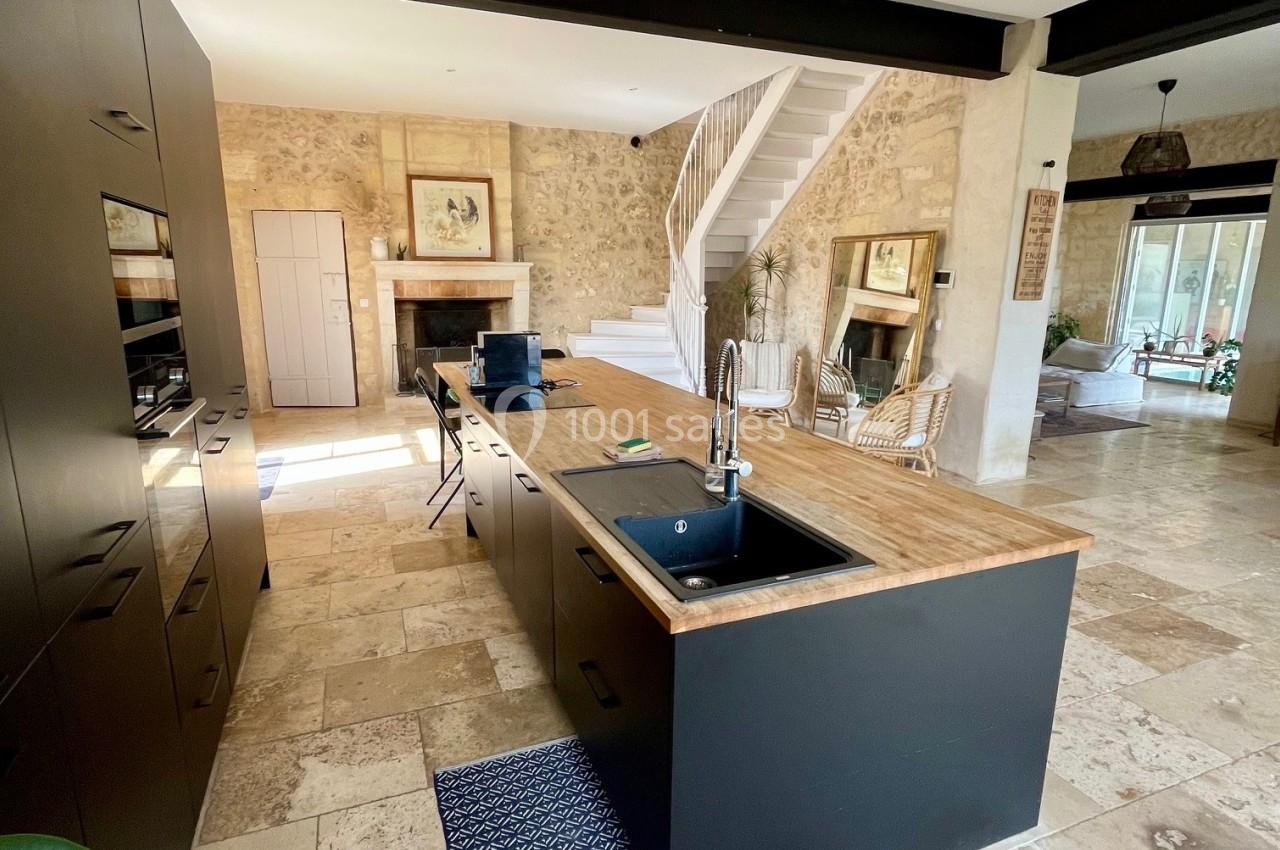 Cuisine moderne avec îlot central en bois, évier intégré, sol en pierre et murs en pierre apparente dans un espace lumineux.