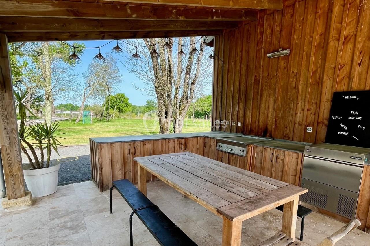 Cuisine d'extérieur en bois avec table, bancs, évier, barbecue intégré et vue sur un jardin arboré.