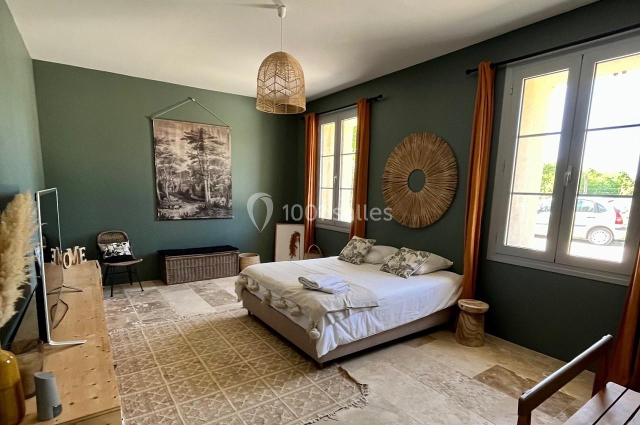 Chambre lumineuse avec lit double, murs verts, décoration naturelle et grandes fenêtres donnant sur l'extérieur.