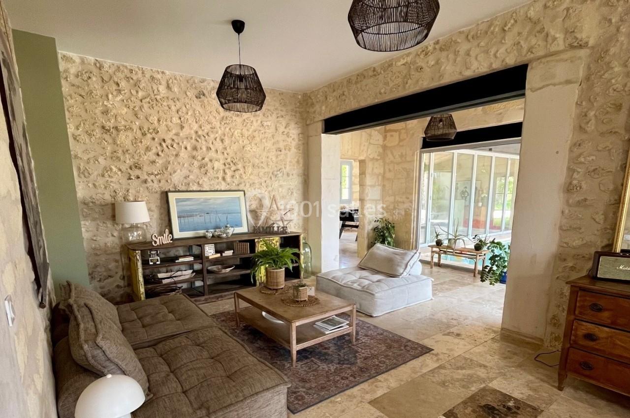 Salon lumineux avec murs en pierre, canapé, table basse, tapis, étagère décorée et vue sur une véranda.