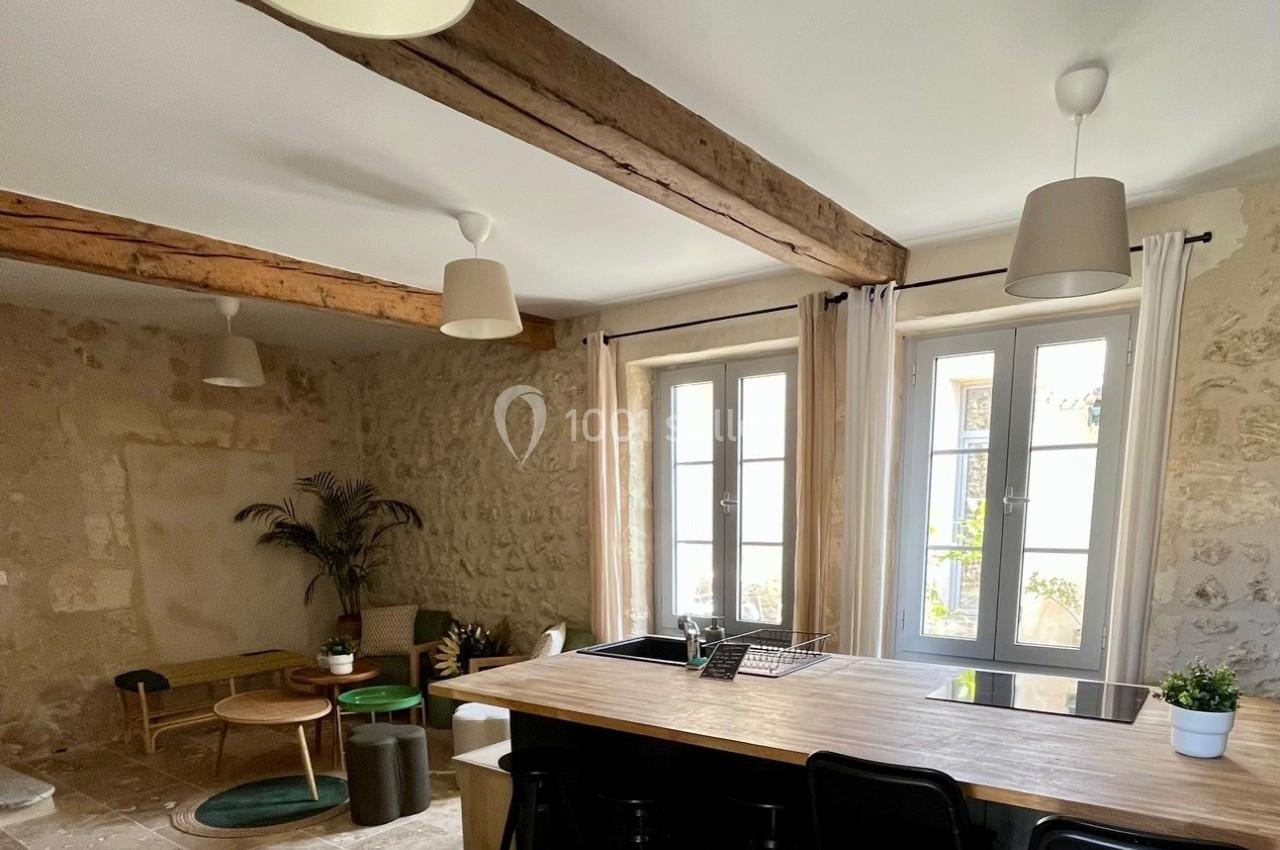 Cuisine ouverte avec îlot central en bois, tabourets noirs, murs en pierre, poutres apparentes et lumière naturelle.