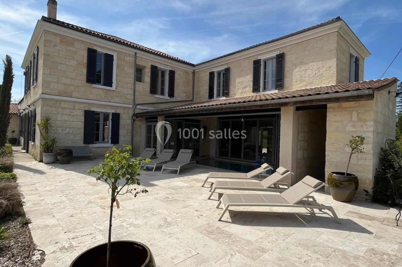 Façade d'une maison en pierre avec terrasse ensoleillée, chaises longues et pots de fleurs sur un sol pavé clair.