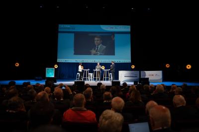 Des personnes échangent dans un espace d'exposition intérieur avec des stands et une structure métallique visible en arrière…