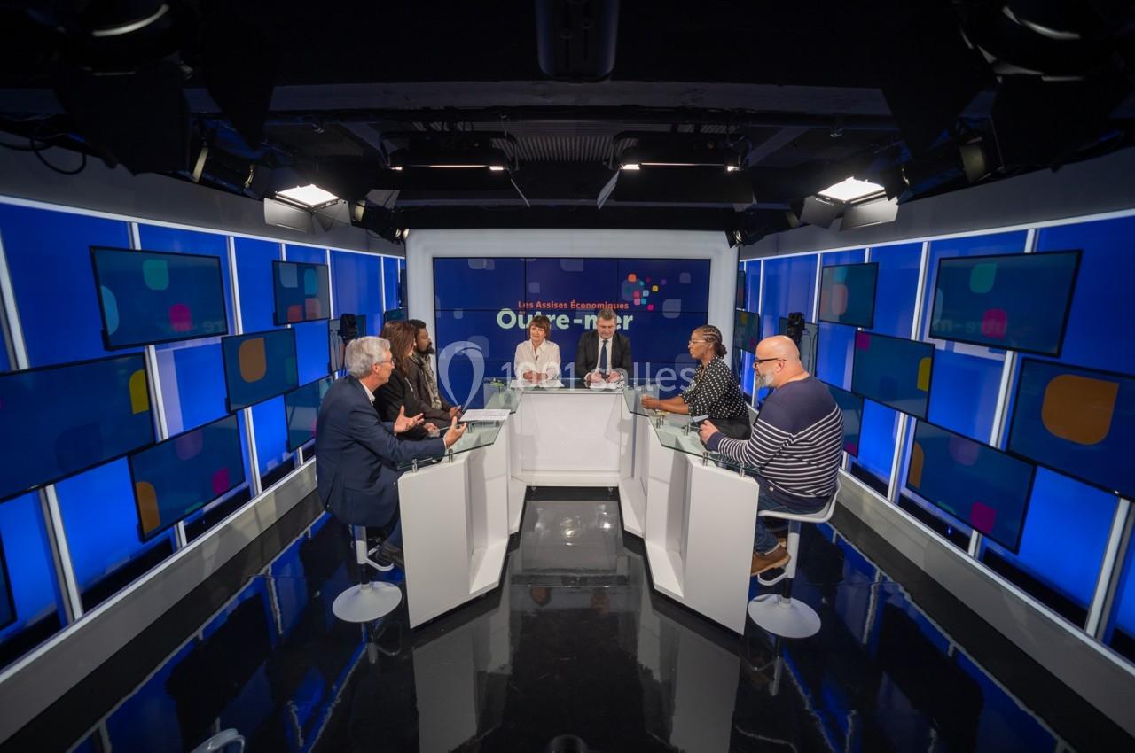 Un groupe de personnes discute autour d'une table dans un studio télévisé éclairé avec des écrans en arrière-plan.