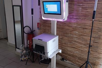 Miniature Le Marchand de Sourires #2 Borne photo sur trépied avec anneaux lumineux, installée dans une salle en briques avec tonneaux en arrière-plan.