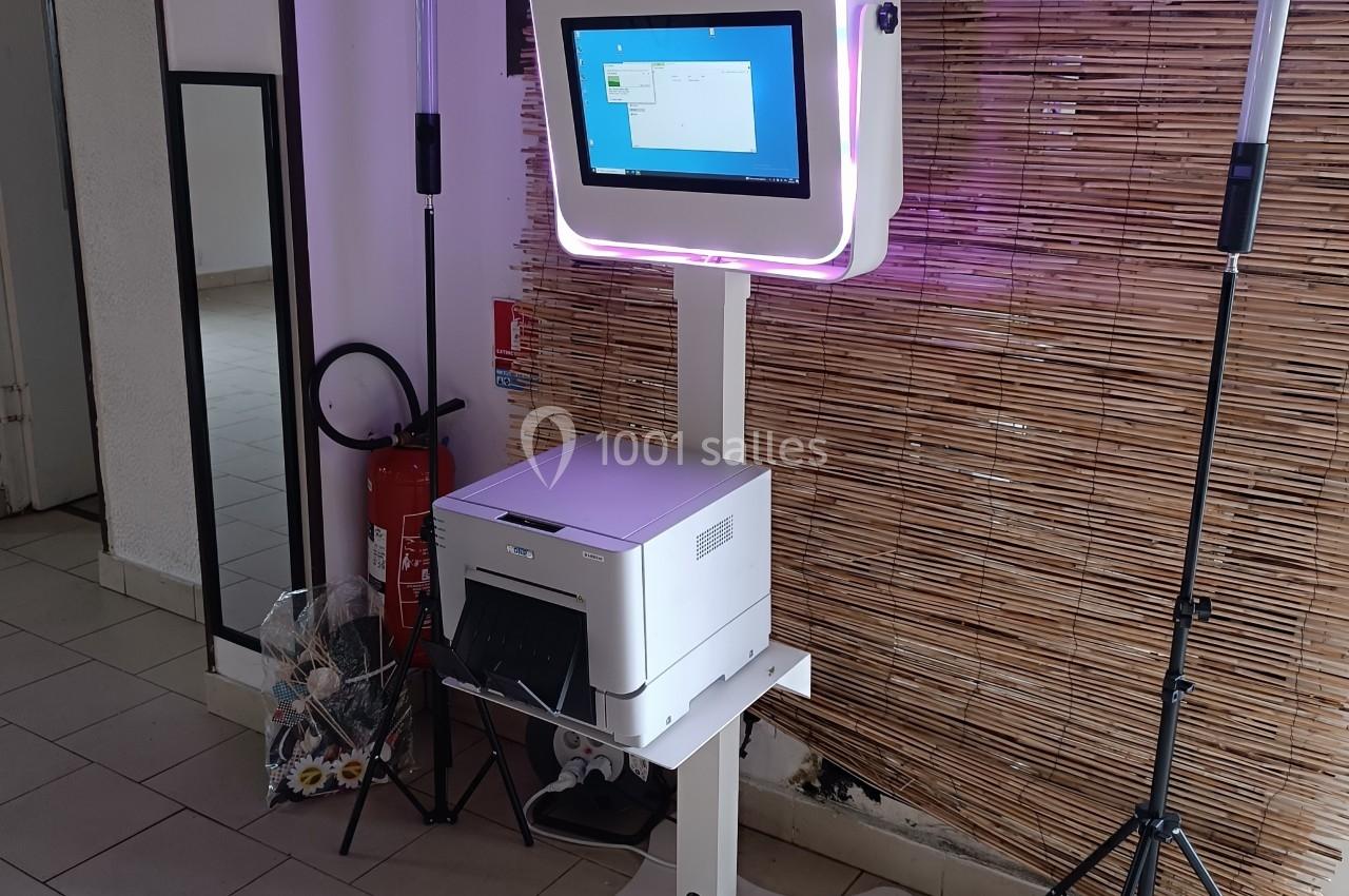 Borne photo interactive avec écran tactile et imprimante, installée devant un mur en bambou dans un espace intérieur.