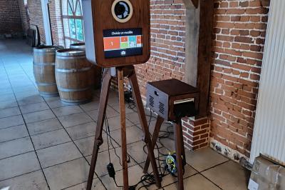 Miniature Le Marchand de Sourires #6 Borne photo sur trépied avec anneaux lumineux, installée dans une salle en briques avec tonneaux en arrière-plan.