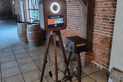 Miniature Le Marchand de Sourires #7 Borne photo sur trépied avec anneaux lumineux, installée dans une salle en briques avec tonneaux en arrière-plan.