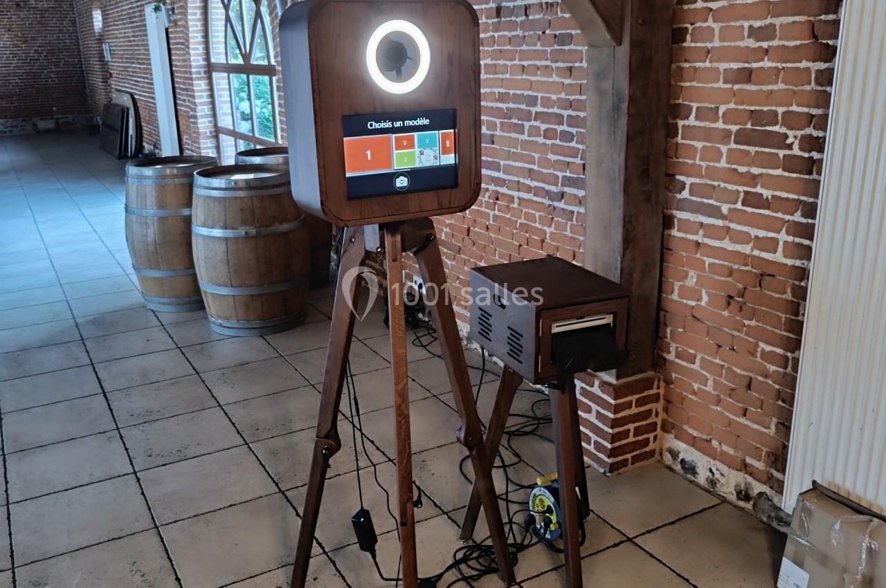 Borne photo sur trépied en bois avec écran tactile et anneau lumineux, installée dans une salle en briques.