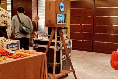 Miniature Le Marchand de Sourires #8 Borne photo sur trépied avec anneaux lumineux, installée dans une salle en briques avec tonneaux en arrière-plan.