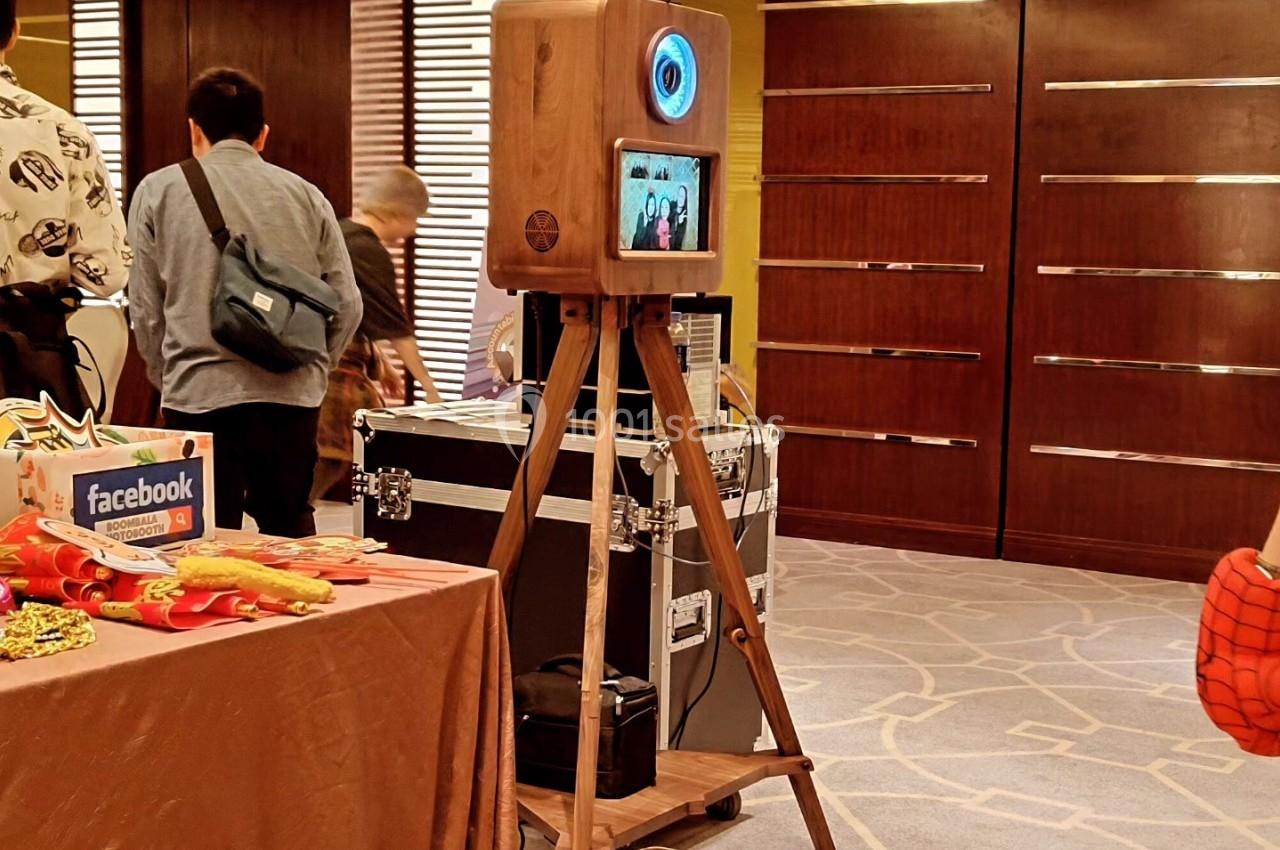 Borne photo en bois sur trépied avec écran et flash, installée dans une salle avec des participants en arrière-plan.