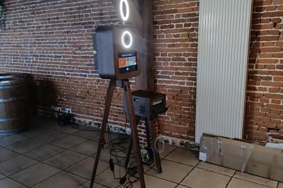 Miniature Le Marchand de Sourires #9 Borne photo sur trépied avec anneaux lumineux, installée dans une salle en briques avec tonneaux en arrière-plan.