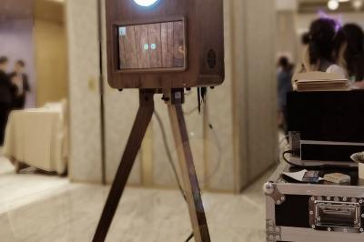 Miniature Le Marchand de Sourires #3 Borne photo sur trépied avec anneaux lumineux, installée dans une salle en briques avec tonneaux en arrière-plan.