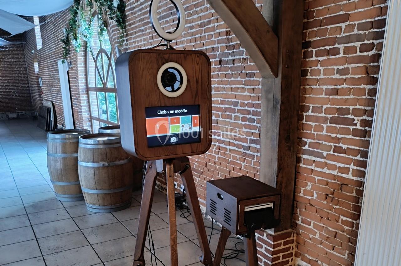 Borne photo vintage en bois sur trépied, installée dans une salle en briques avec des tonneaux en arrière-plan.