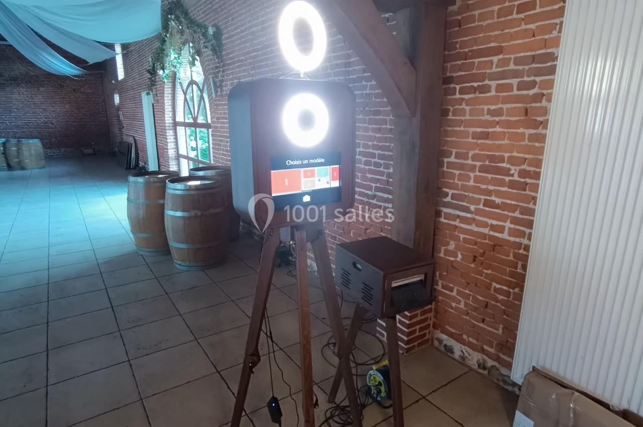 Borne photo sur trépied avec éclairage circulaire, installée dans une salle en briques avec tonneaux en arrière-plan.