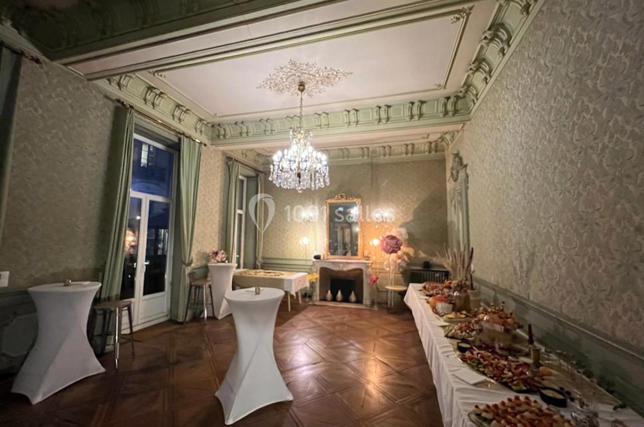 Salle élégante avec moulures, lustre, tables hautes et buffet garni sur des nappes blanches.