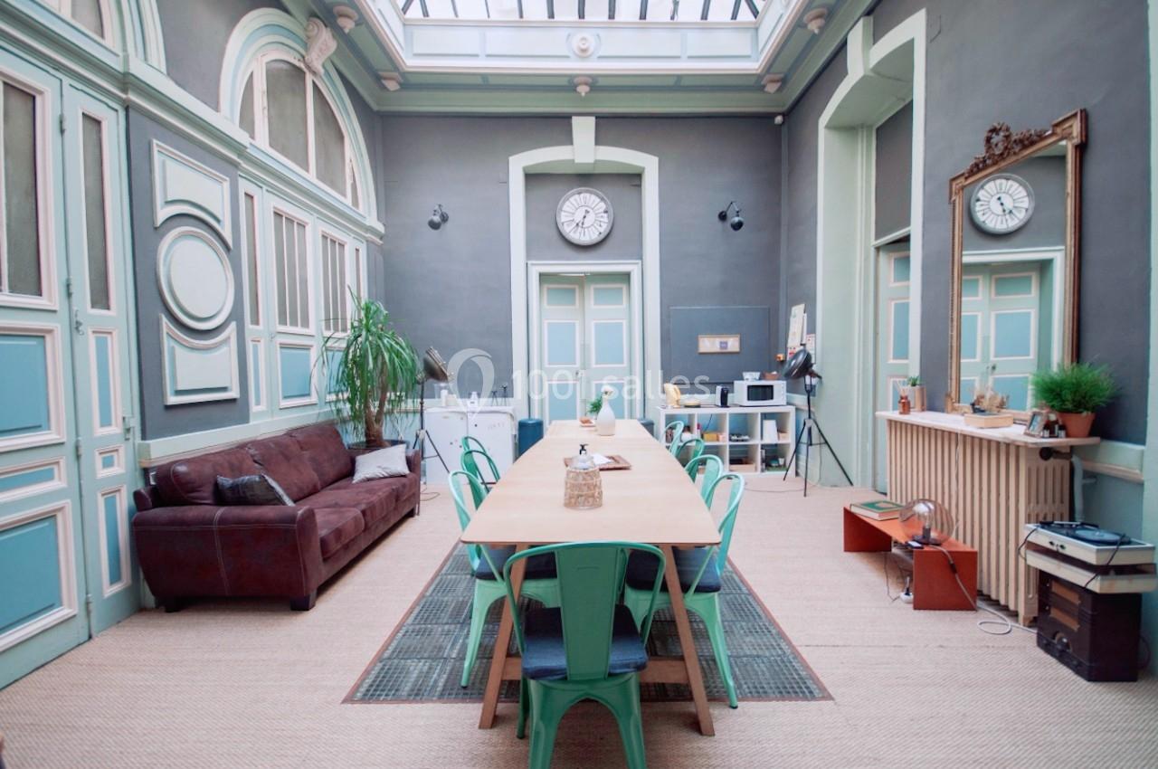 Salle lumineuse avec grande table en bois entourée de chaises vertes, canapé en cuir, plantes et décoration vintage.