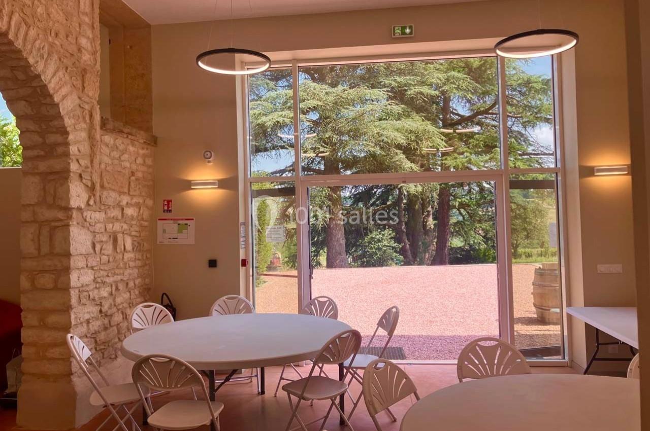 Salle lumineuse avec des tables rondes et des chaises blanches, vue sur un extérieur arboré à travers une grande baie vitrée.