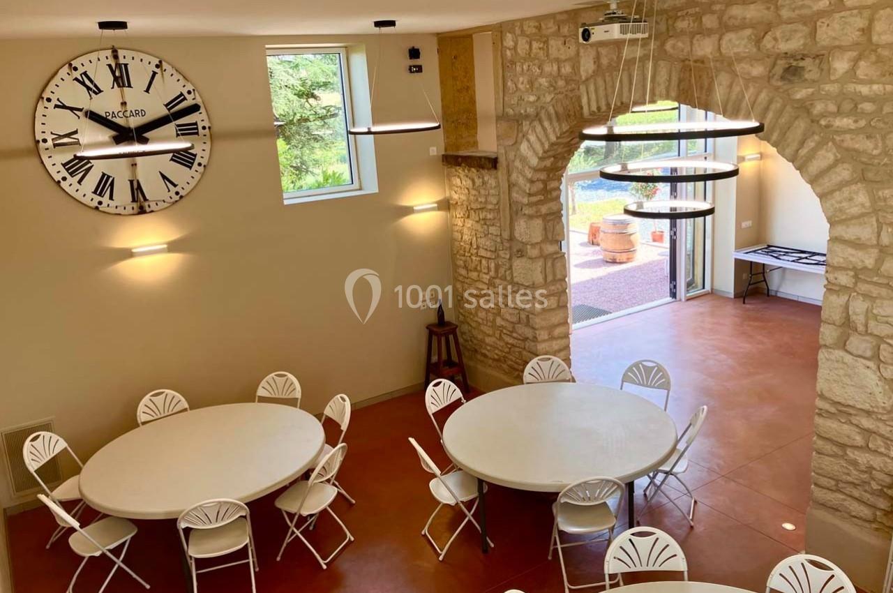 Salle lumineuse avec murs en pierre, tables rondes blanches, chaises pliantes et vue sur un espace extérieur.