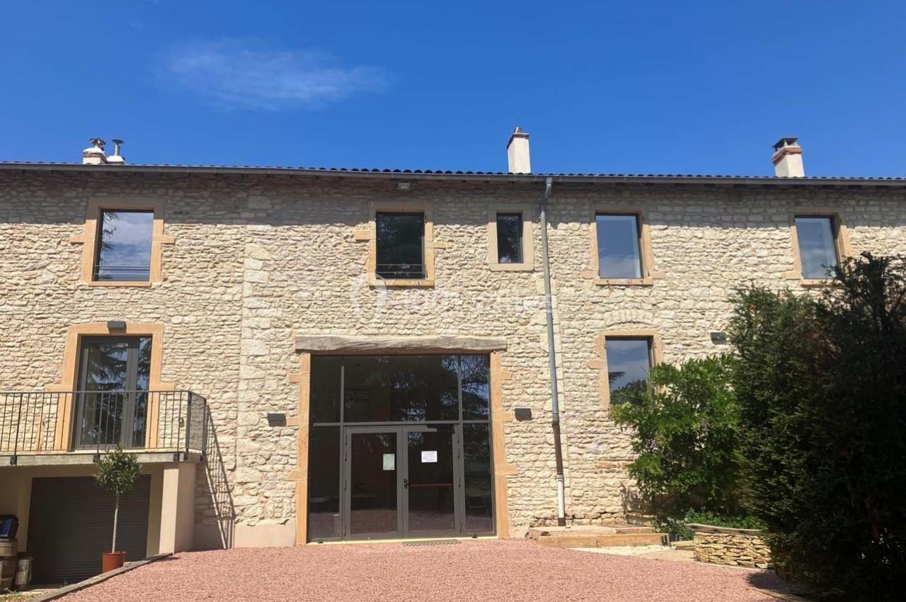 Façade d'un bâtiment en pierre avec de grandes fenêtres, une entrée vitrée et une cour en gravier sous un ciel bleu.