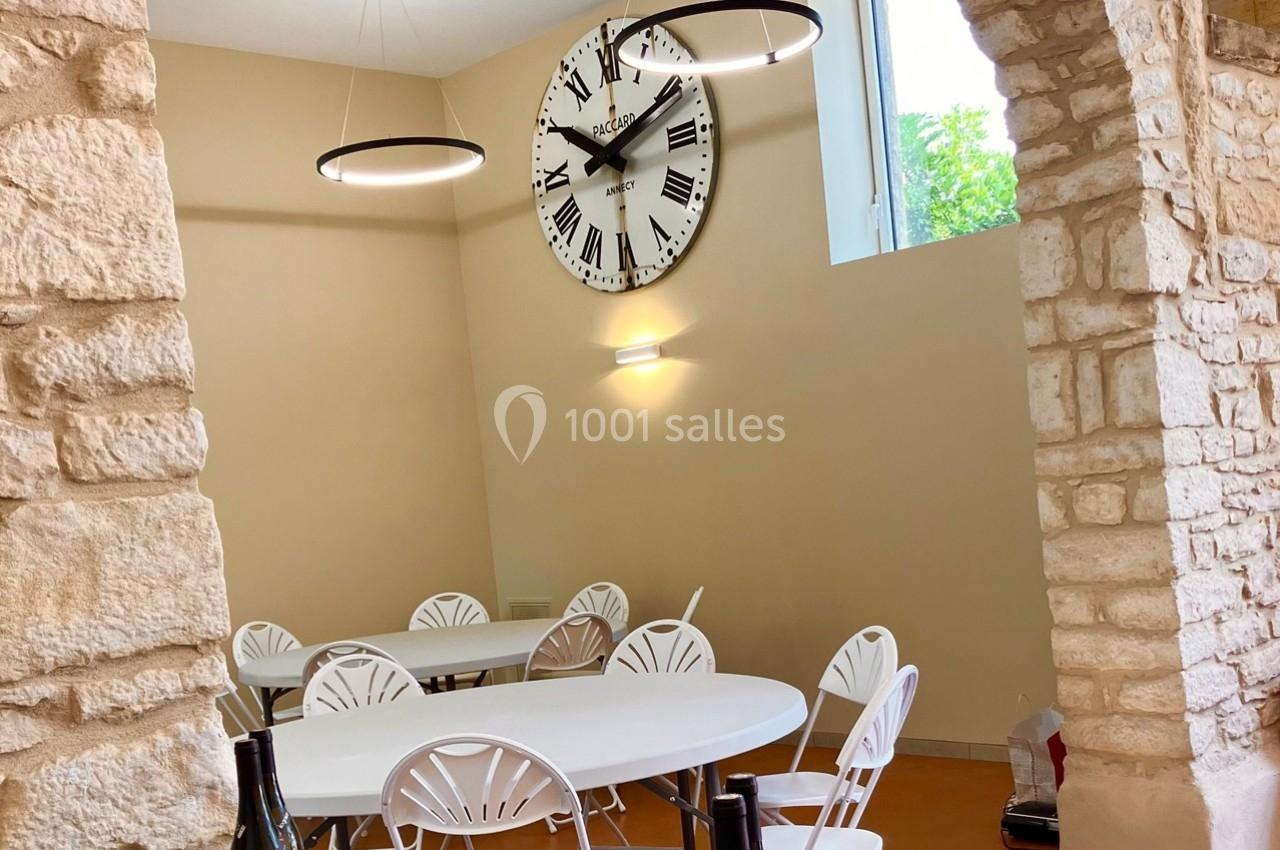 Salle lumineuse avec des murs en pierre, des tables rondes blanches, des chaises assorties et une grande horloge murale.