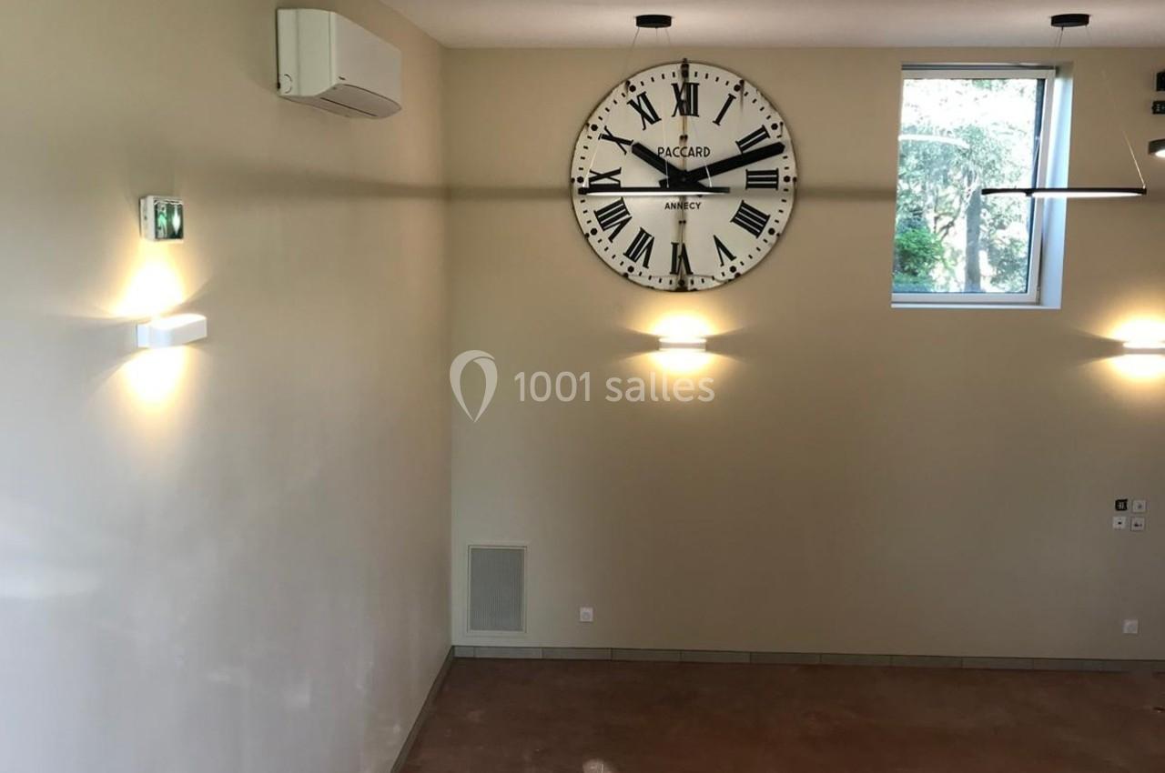 Horloge murale noire et blanche sur un mur beige, éclairée par des appliques, dans une pièce lumineuse avec fenêtre.