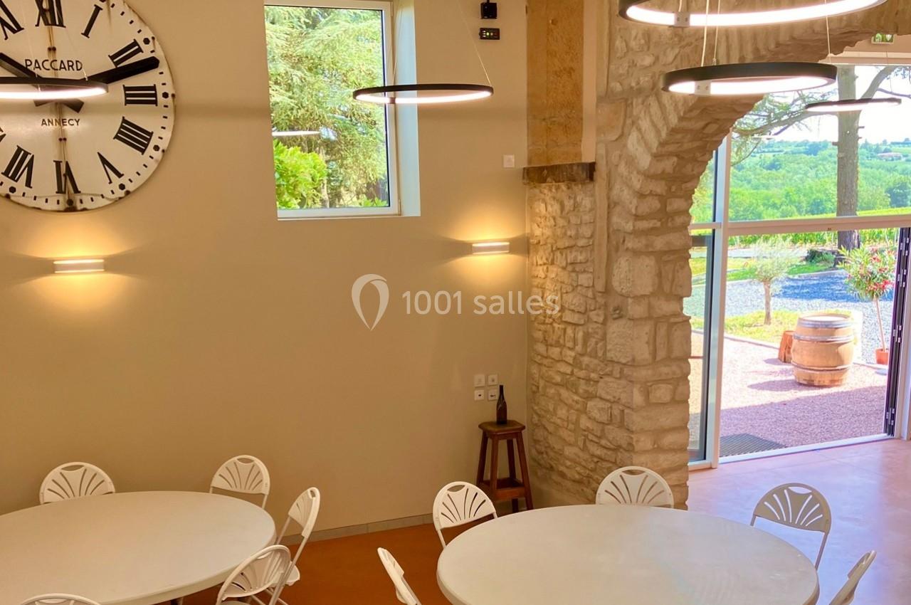 Salle lumineuse avec murs en pierre, tables rondes blanches, grande horloge murale et vue sur un paysage verdoyant.