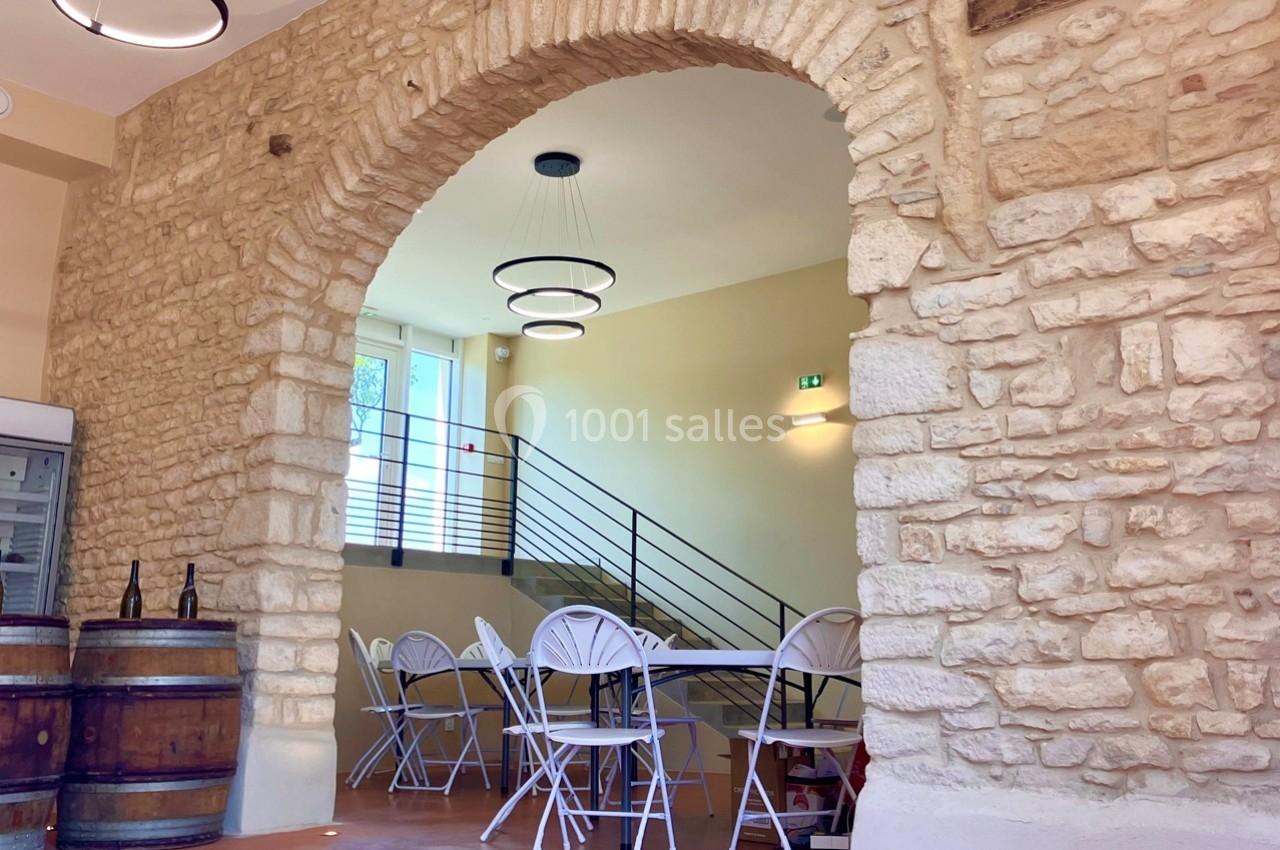 Salle lumineuse avec murs en pierre, tables et chaises blanches, barils en bois et escalier en arrière-plan.