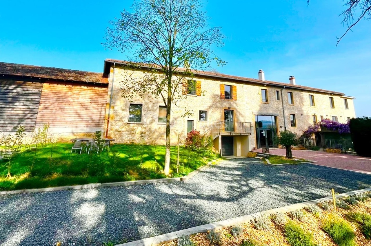 Bâtiment en pierre rénové avec un jardin, une allée en gravier et des arbres sous un ciel ensoleillé.
