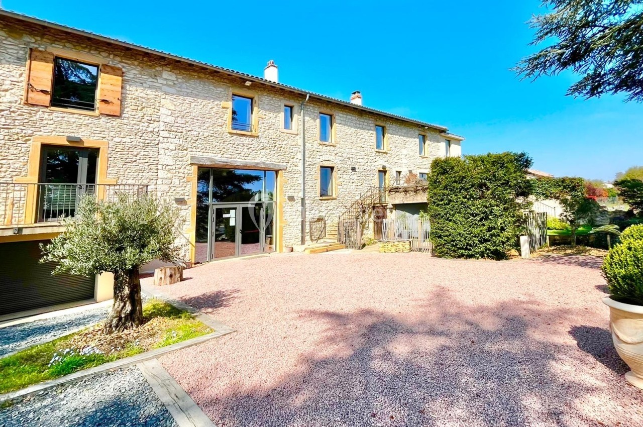 Façade d'une maison en pierre avec volets en bois, grande cour gravillonnée et végétation environnante.