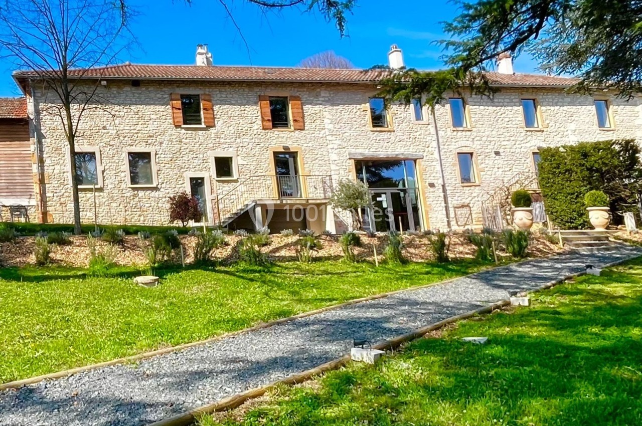 Bâtisse en pierre avec grandes fenêtres, entourée d'un jardin verdoyant et d'un chemin gravillonné.