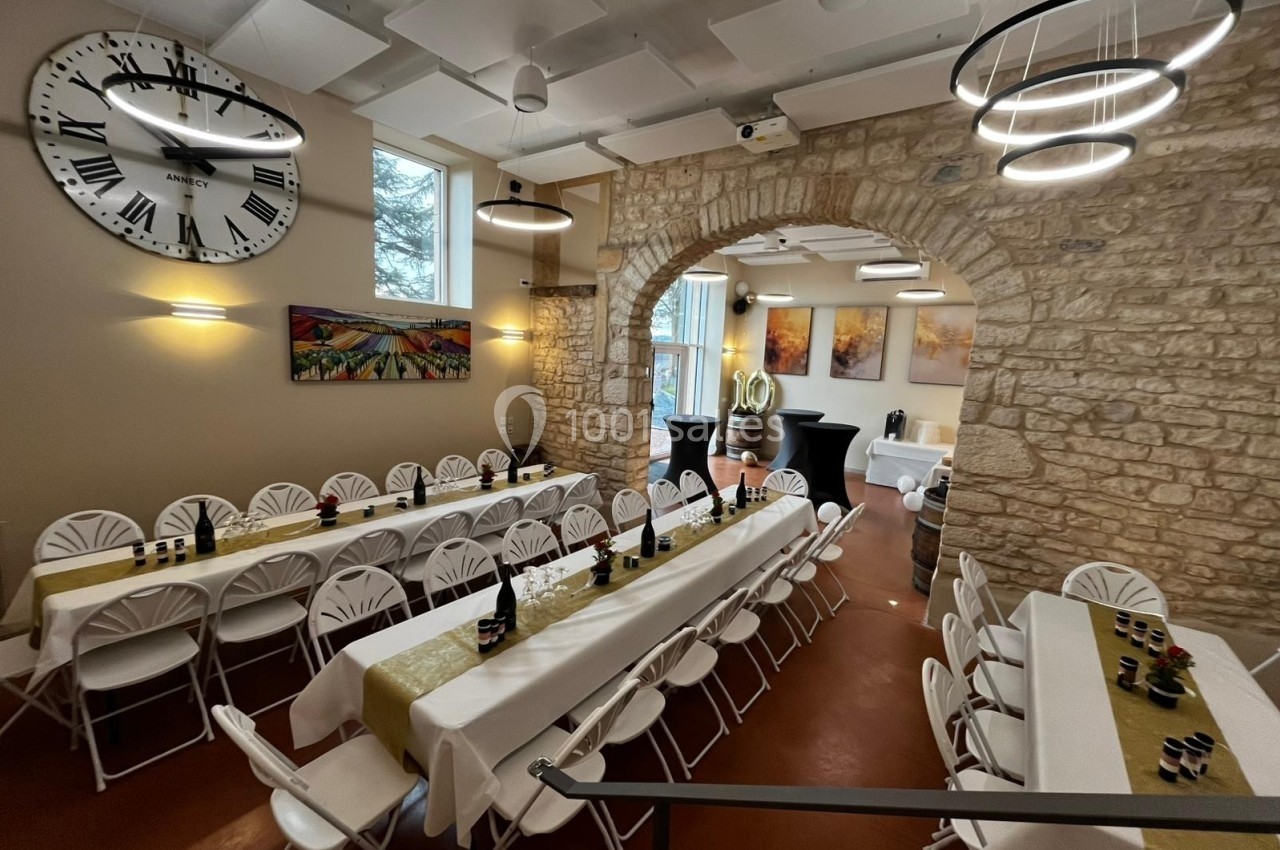 Salle lumineuse avec murs en pierre, tables décorées pour un événement, grandes horloges et éclairage moderne.