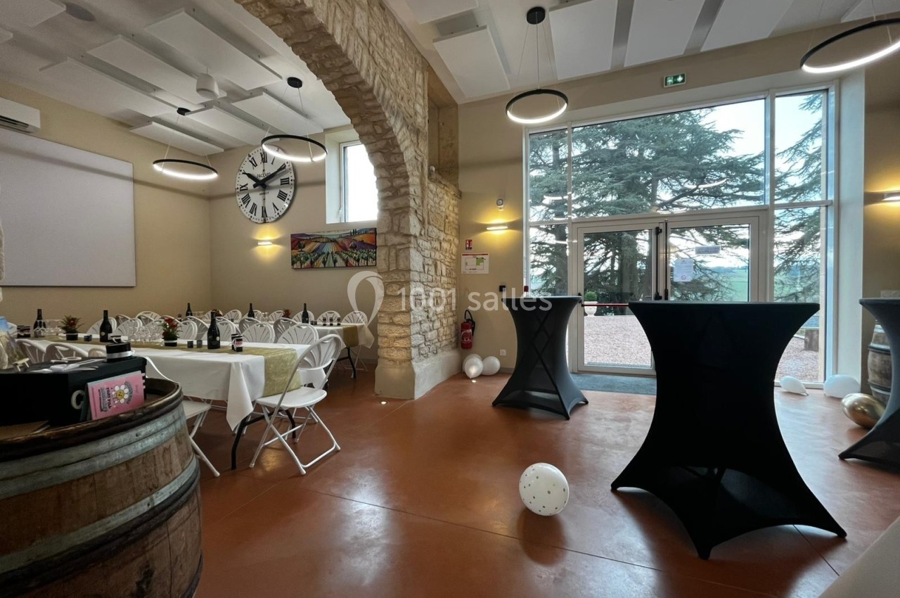 Salle de réception avec tables dressées, tonneau en premier plan, grandes baies vitrées et vue sur des arbres.