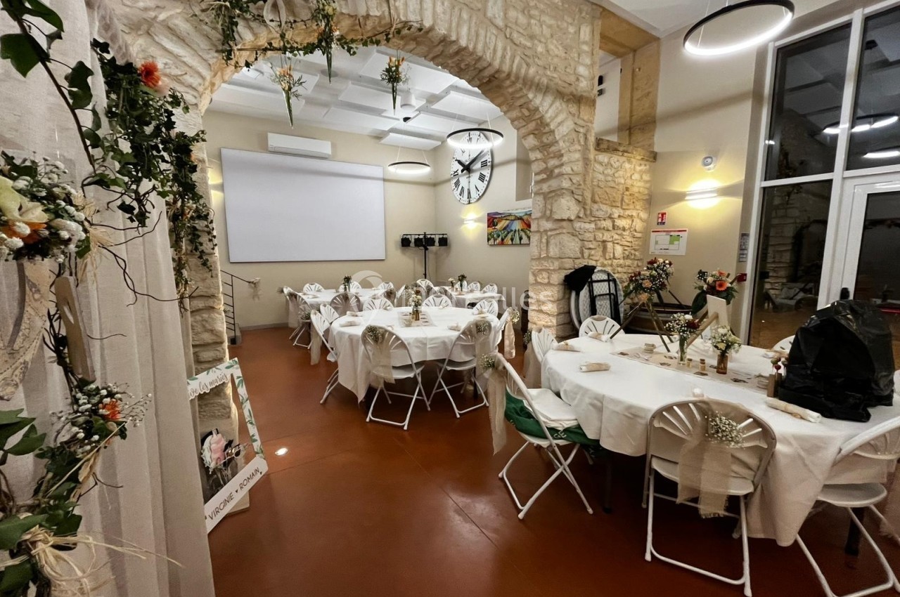 Salle décorée pour un événement, avec tables rondes dressées, chaises blanches et arches en pierre ornées de fleurs.