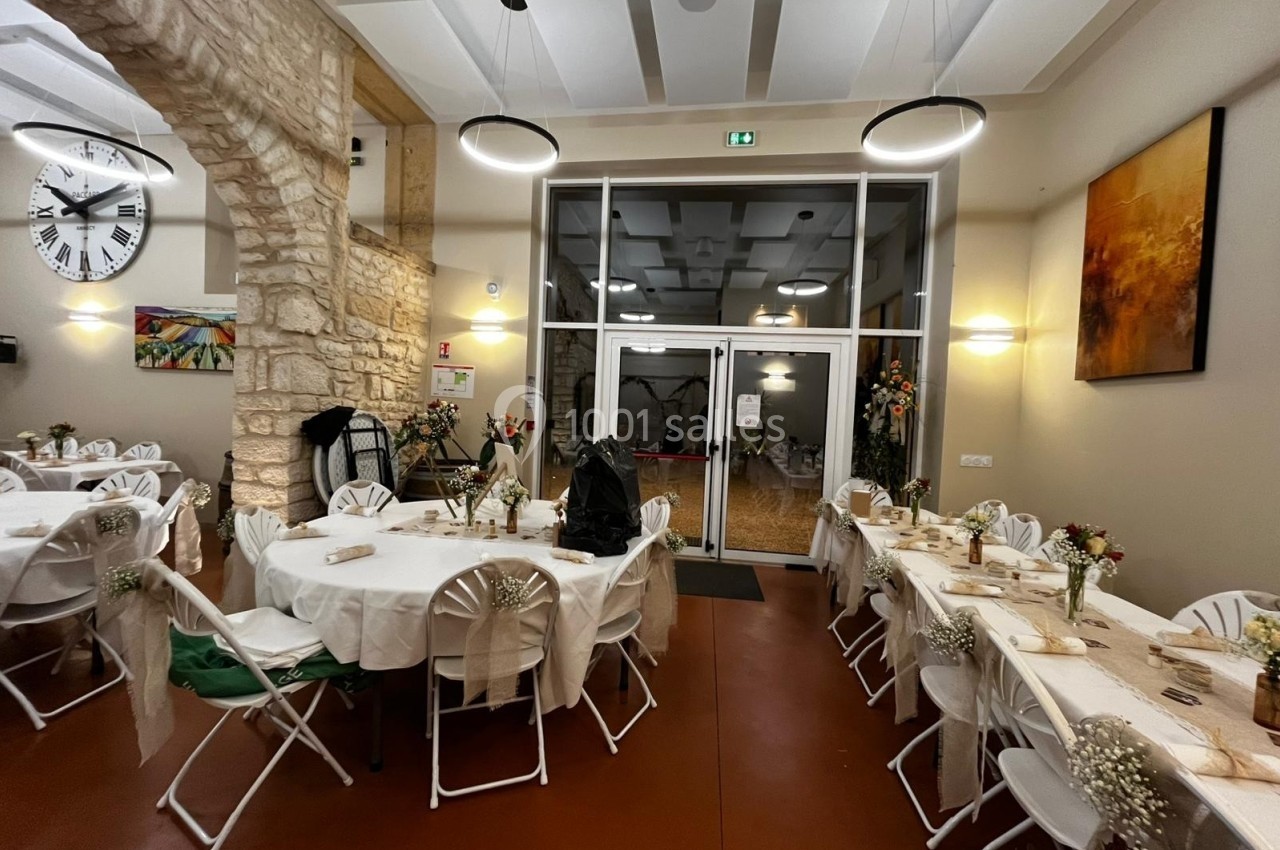 Salle décorée pour un événement, avec des tables rondes dressées, des chaises blanches et des fleurs en centre de table.
