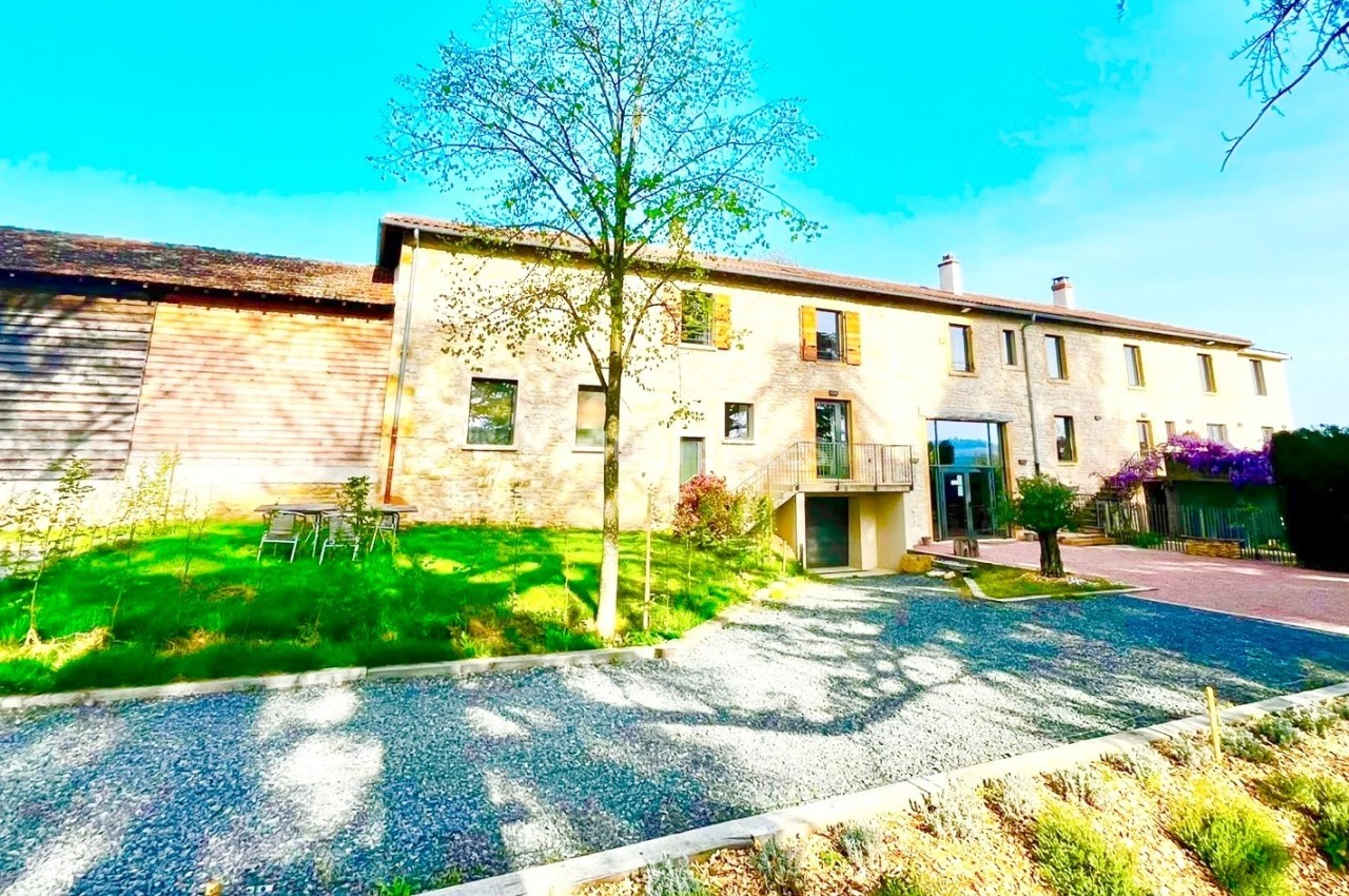 Bâtiment en pierre avec un jardin, une allée gravillonnée et des arbres sous un ciel dégagé.