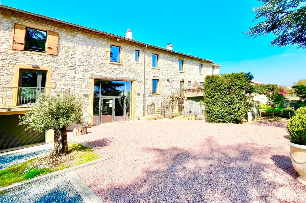 Façade en pierre d'une grande maison avec volets en bois, entourée d'une cour gravillonnée et de végétation.
