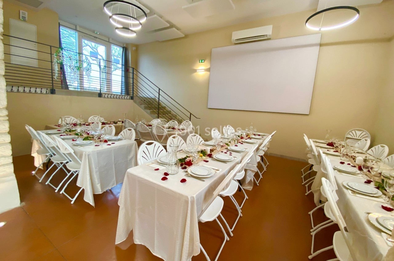 Salle de réception lumineuse avec tables dressées, nappes blanches, chaises pliantes et décoration florale.