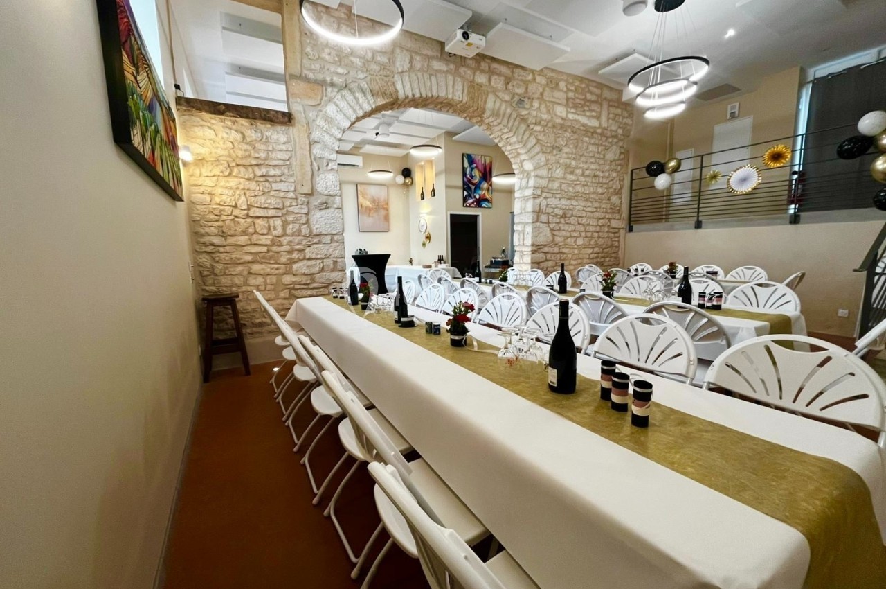 Salle décorée avec des tables longues, nappes blanches, chaises alignées et murs en pierre apparente.