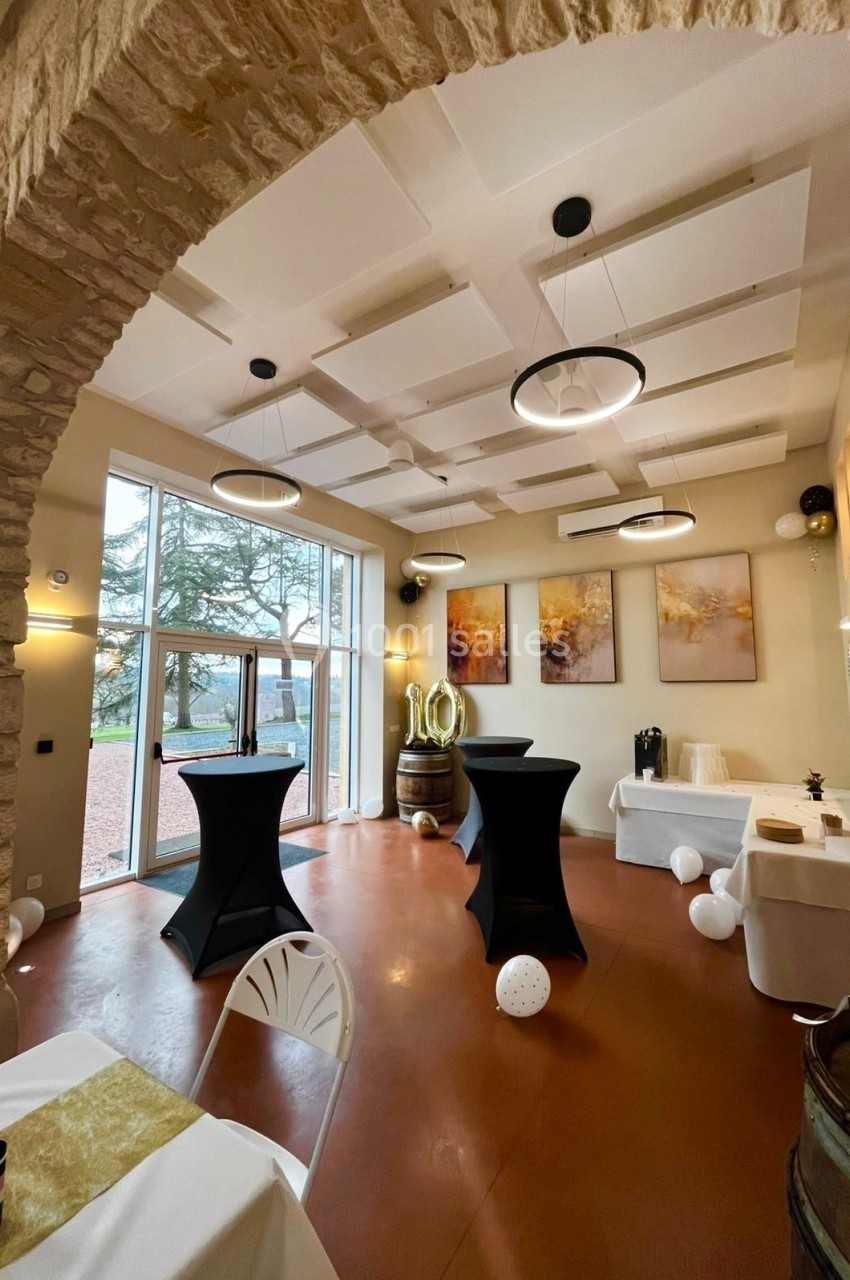 Salle décorée pour un événement, avec tables hautes, ballons, grandes fenêtres et peintures murales.